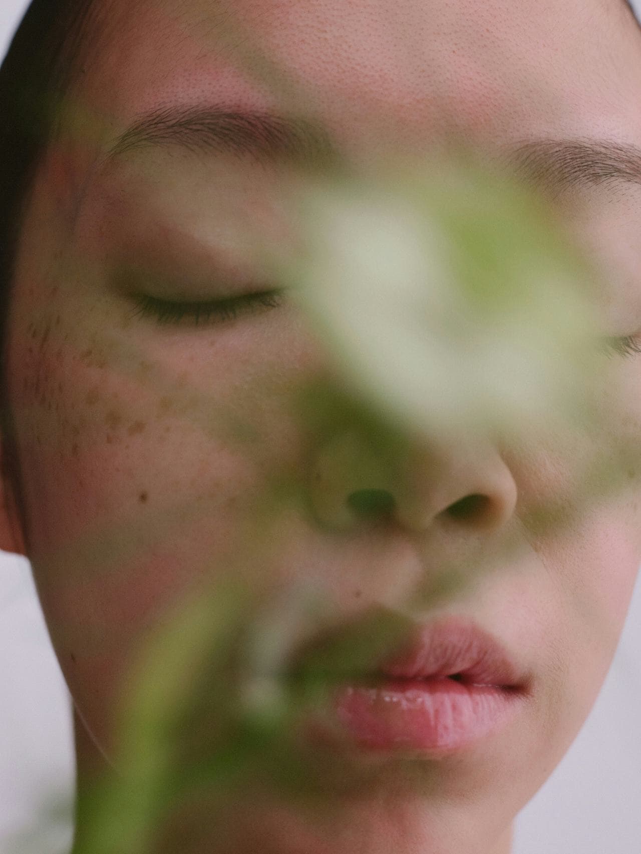 Close-up of a woman's contemplative face with eyes closed, partially obscured by a plant, symbolizing reflection and healing after a cancer diagnosis.